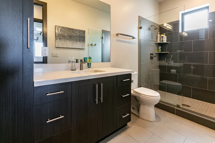 Bathroom featuring a dark wood-finish vanity with a light countertop, a glass-enclosed shower with dark rectangular wall tiles, and light-colored floor tiles