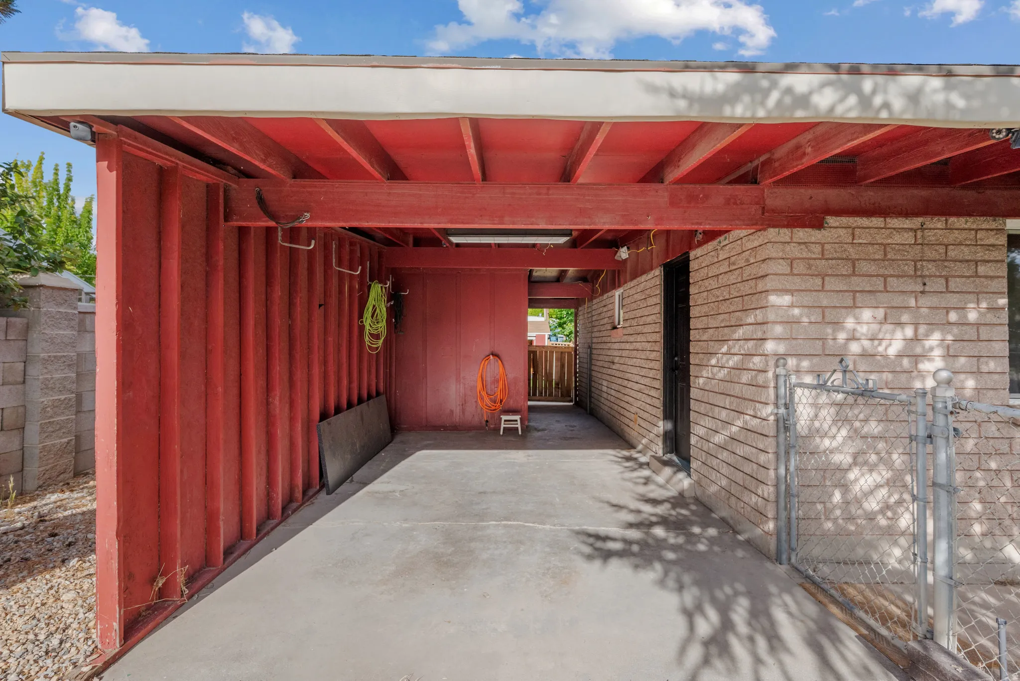 Covered carport with a concrete floor, exposed red ceiling beams, and a light-colored brick exterior wall