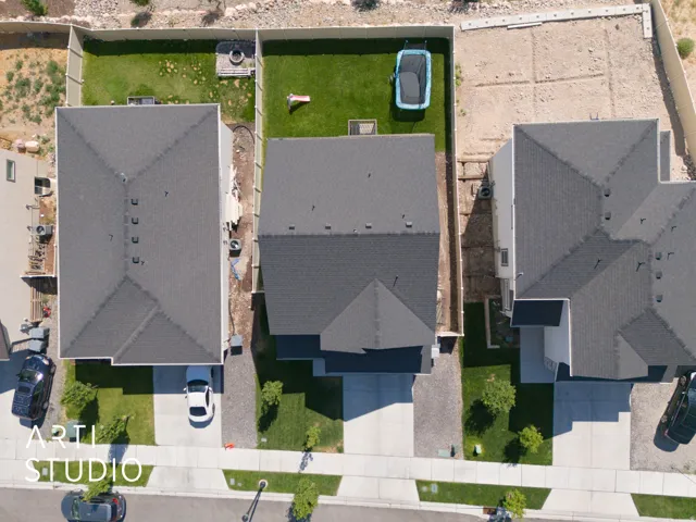 Aerial perspective showcasing a property with a dark shingle roof, a light-colored driveway, and a partially landscaped backyard featuring a green lawn