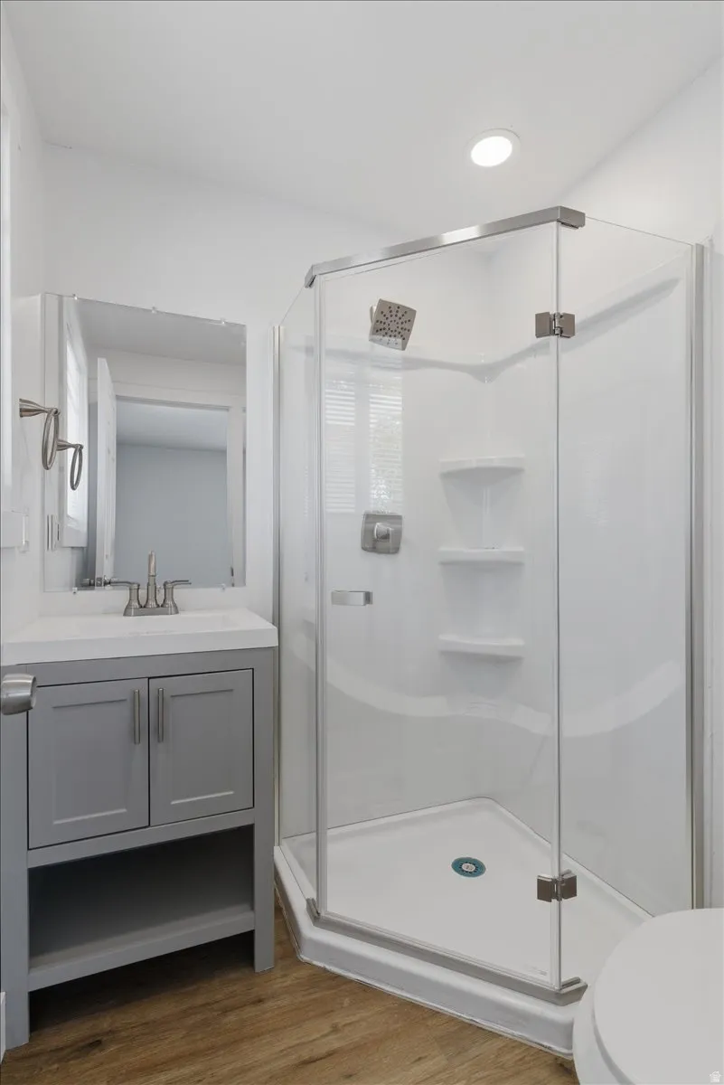 Modern bathroom featuring a glass-enclosed corner shower with built-in shelving, a gray vanity with a white countertop and integrated sink, brushed nickel fixtures, recessed lighting, and wood-finish flooring