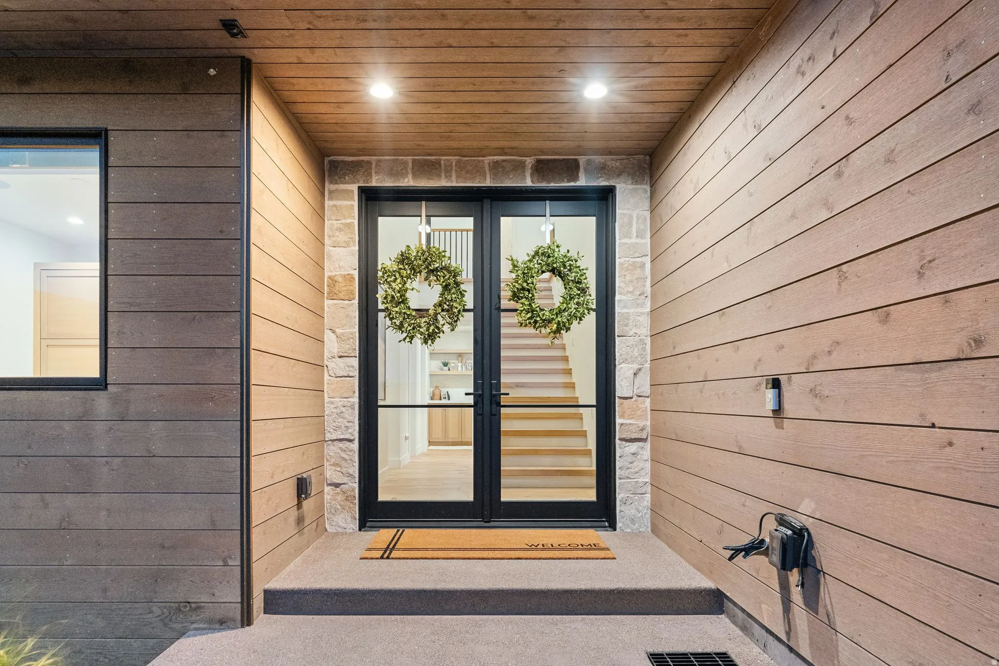 Modern architectural entrance featuring dark-framed double glass doors, stone facade accents, horizontal wood-finish siding, and a wood-panelled ceiling with recessed lighting