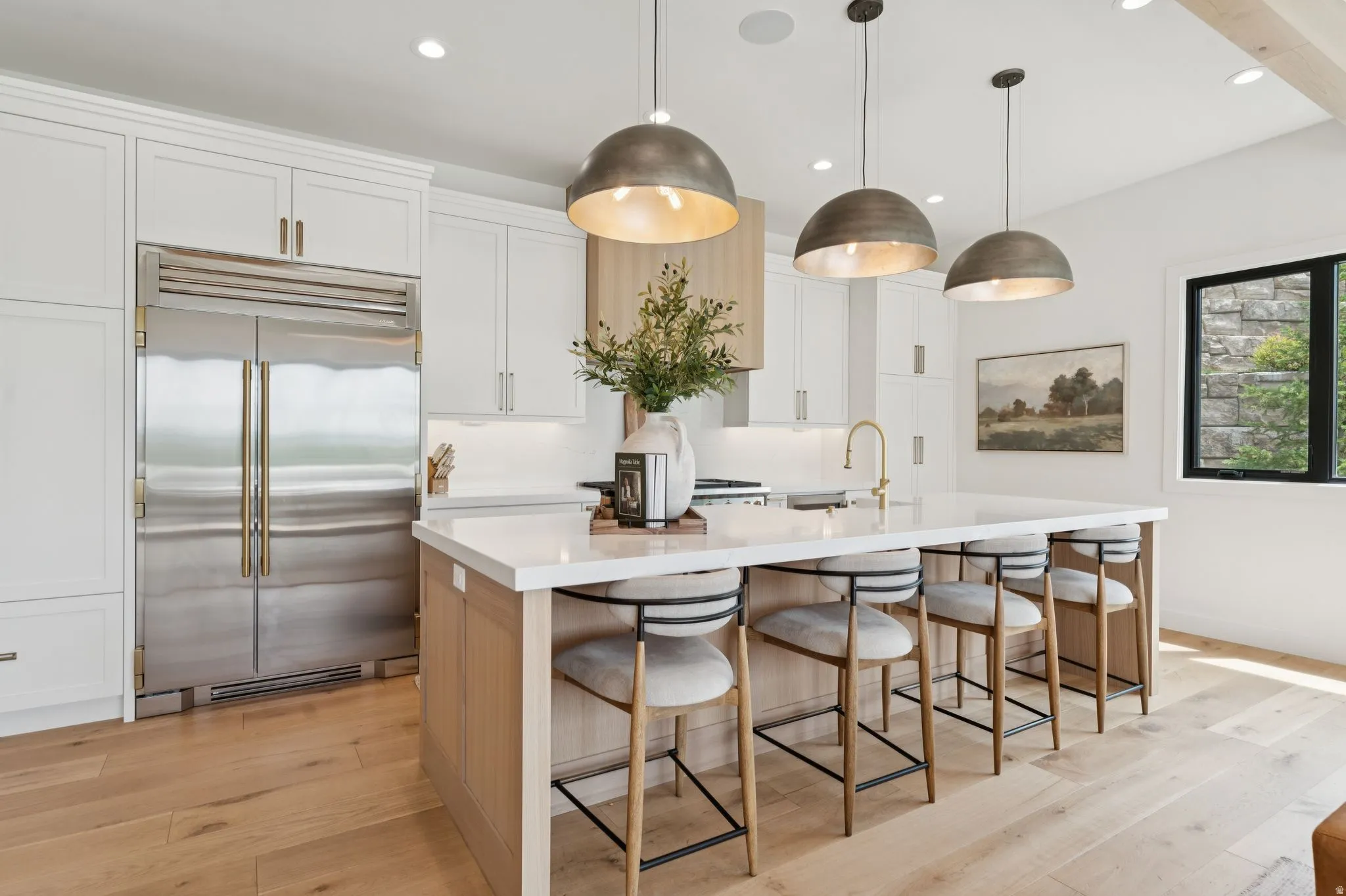 Gourmet kitchen featuring a large central island with seating, a stainless steel built-in refrigerator, light wood-finish flooring, white cabinetry with gold-tone hardware, and three dome pendant lights