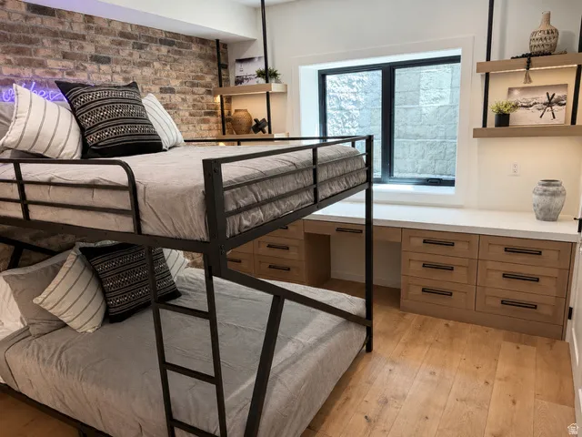 Room featuring a full brick accent wall, wood-finish flooring, a built-in window desk with light wood cabinetry, white countertops, and open shelving with integrated lighting