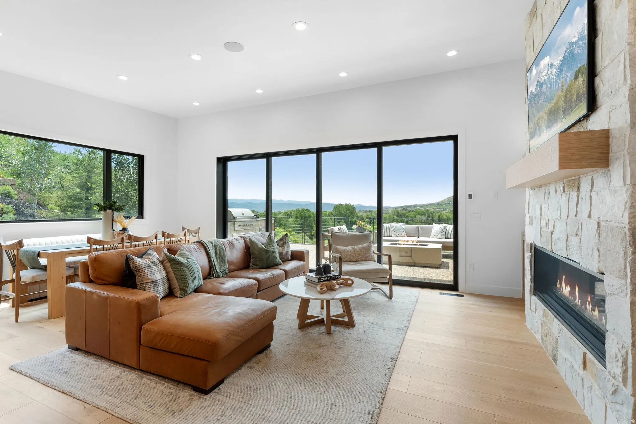 Spacious living area featuring a stone-clad fireplace with a built-in mantel, expansive sliding glass doors, large picture windows, recessed lighting, and light wood-finish flooring