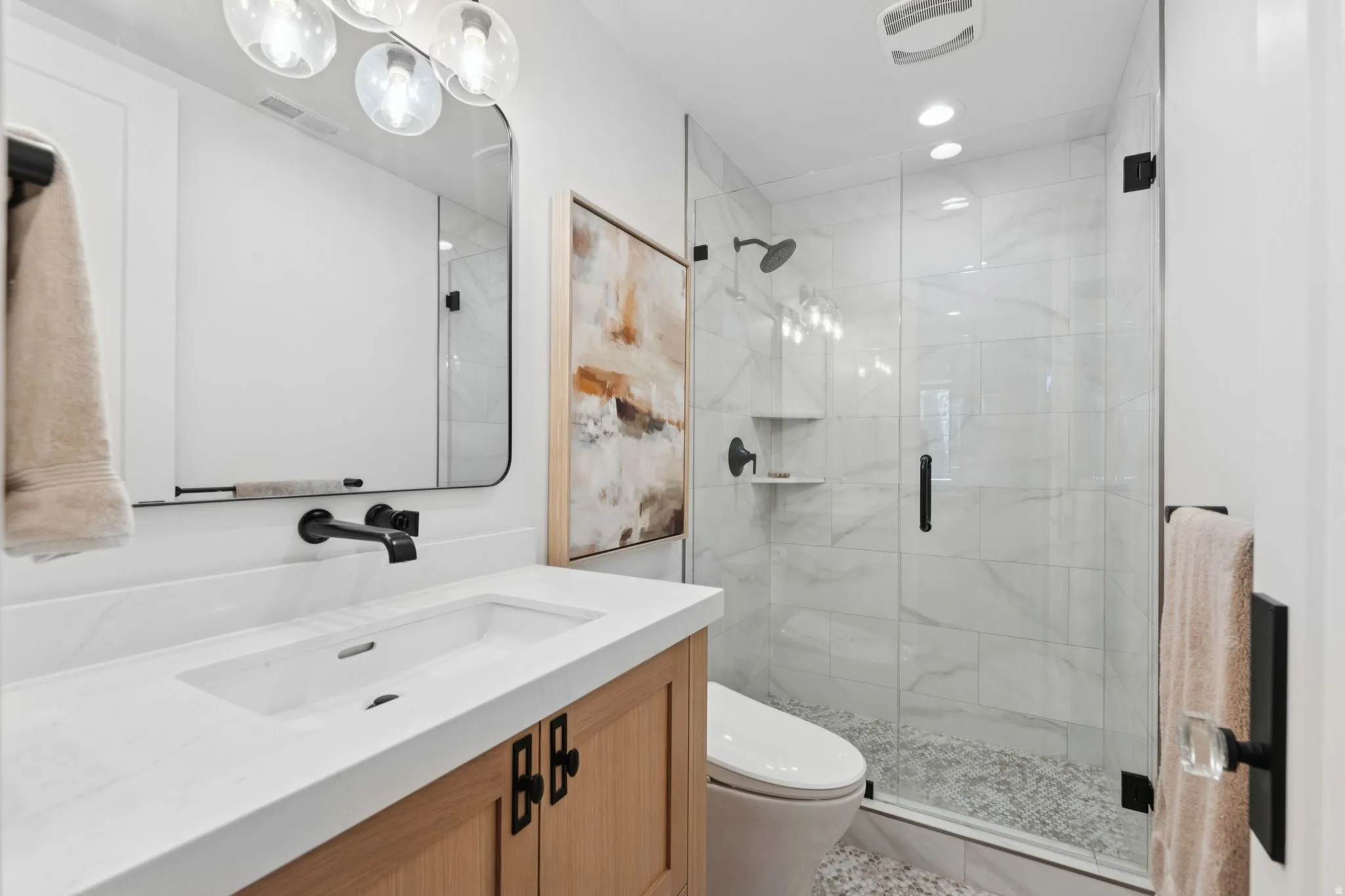 Bathroom featuring a wall-mounted faucet, quartz countertop, wood-finish vanity, glass-enclosed shower with tile surround, and pebbled shower flooring