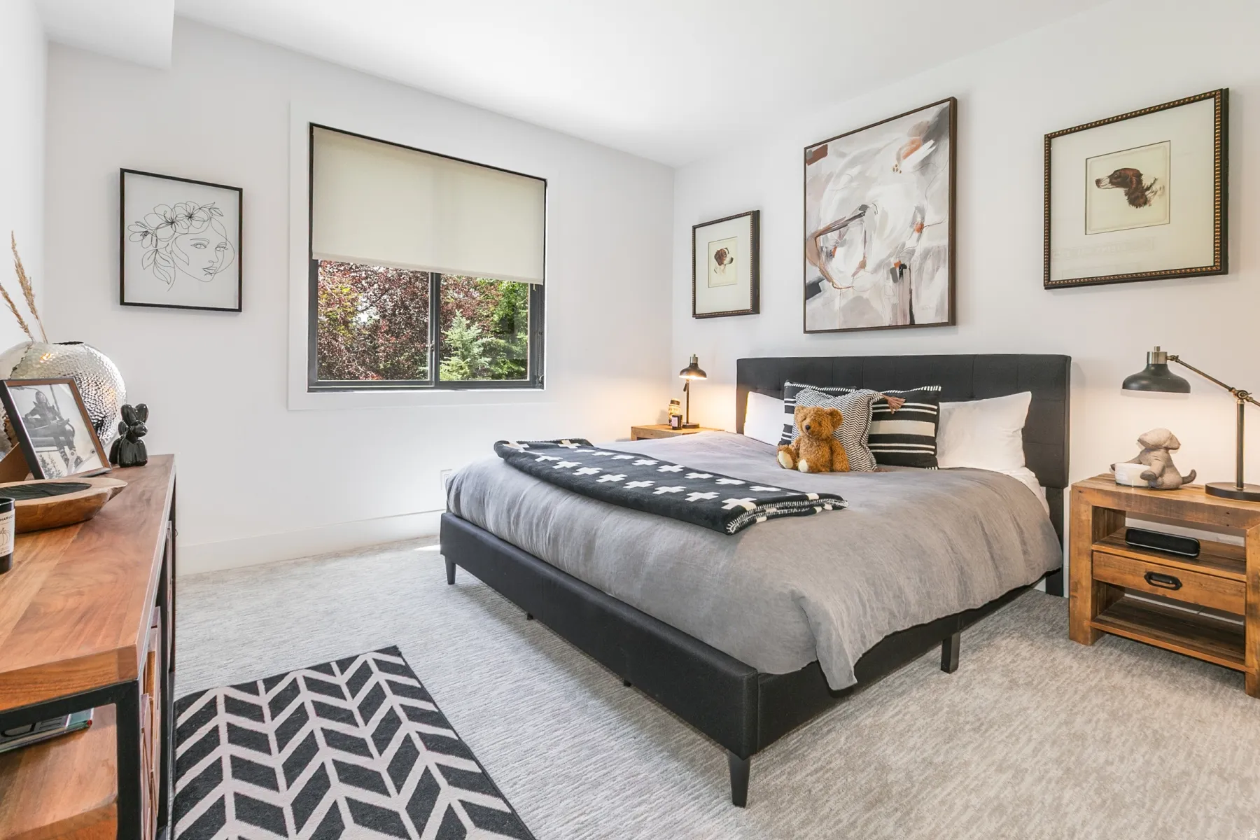 Carpeted room featuring a window with roller shade, a dark upholstered bed frame, and wood-finish nightstands