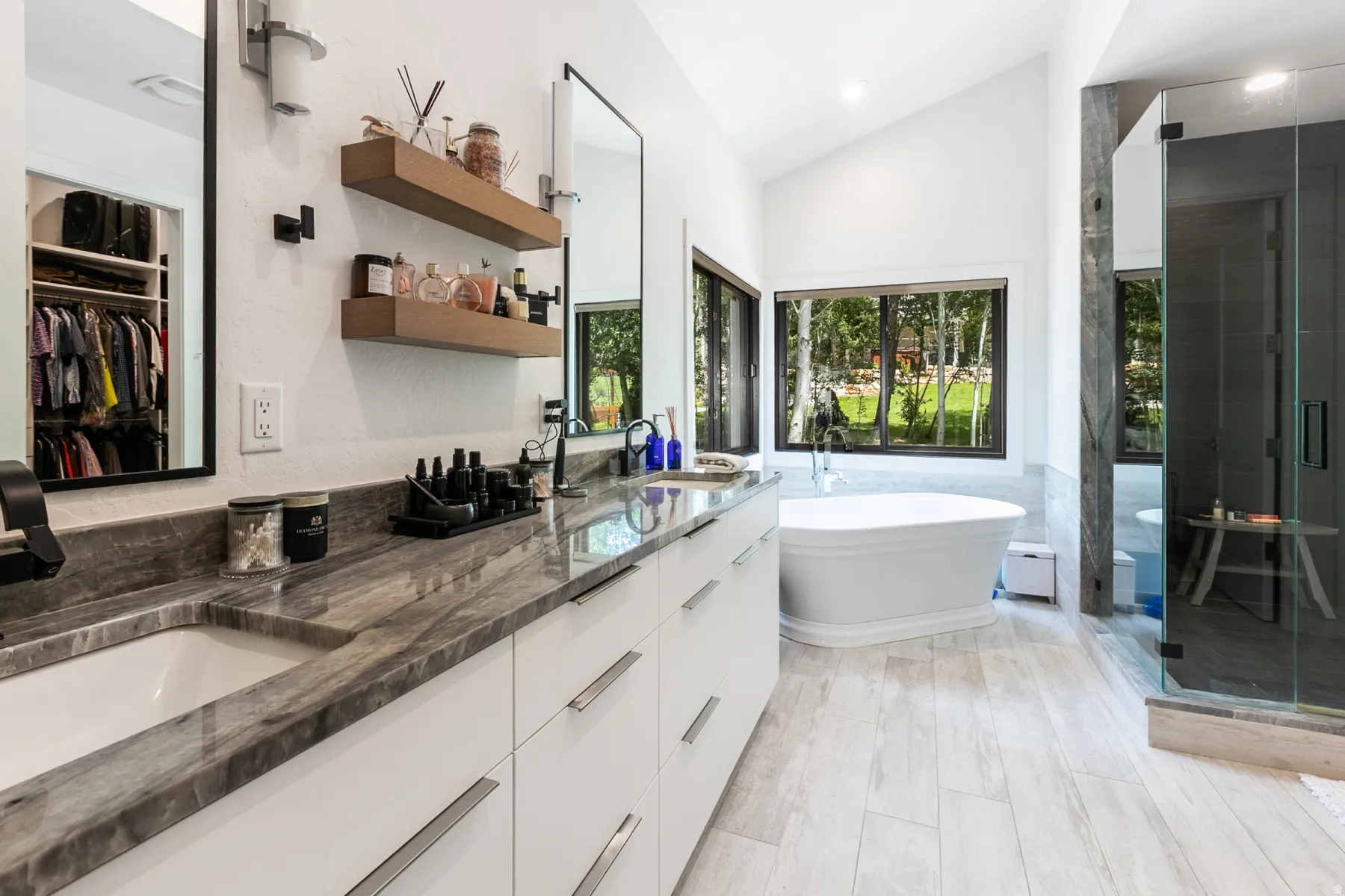 Spacious bathroom featuring a double vanity with stone countertops, a freestanding soaking tub, a glass-enclosed shower, wood-finish flooring, and ample natural light through large windows