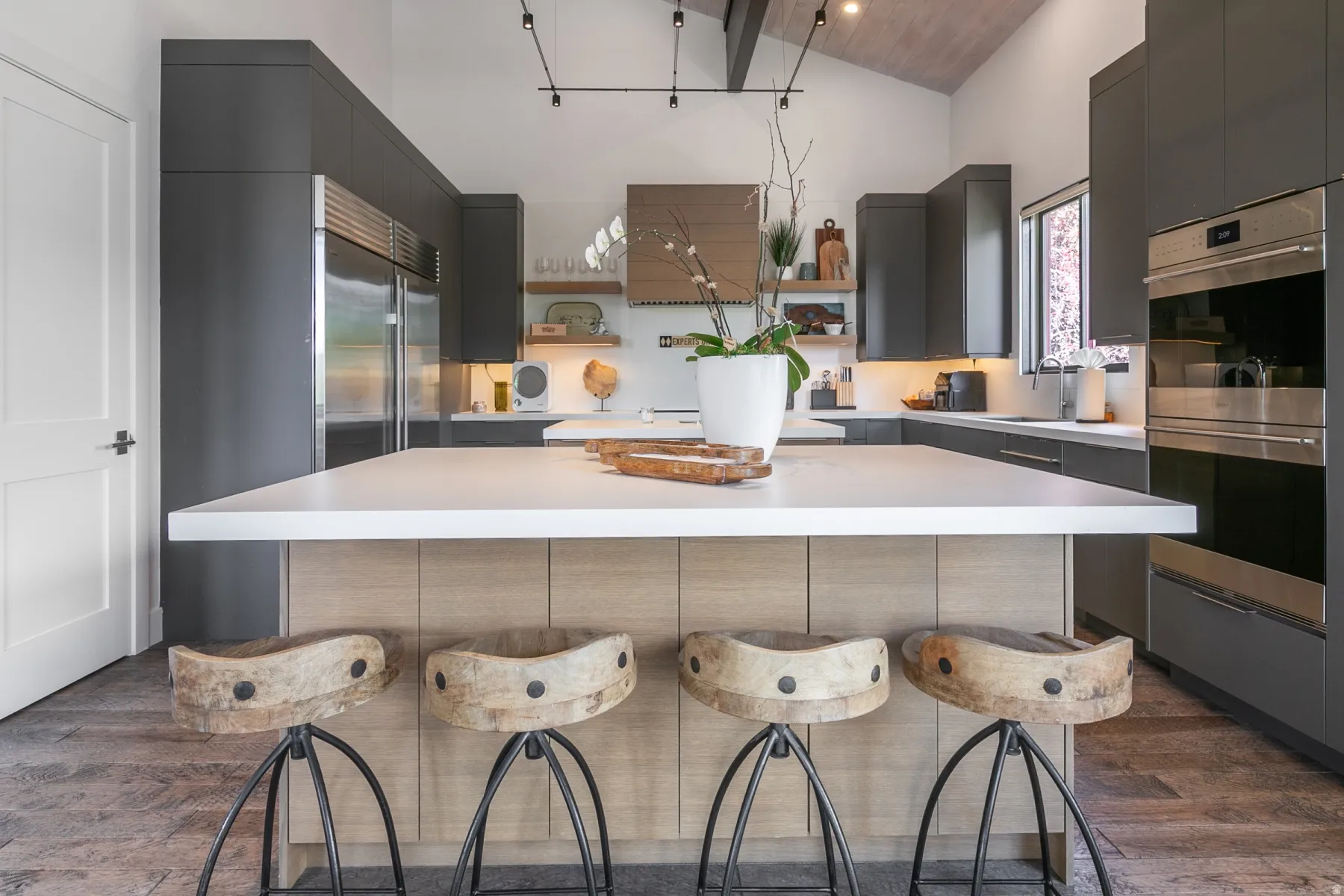 Gourmet kitchen featuring a large central island with light wood-finish cabinetry, sleek dark gray upper and lower cabinetry, stainless steel built-in double ovens, and a stainless steel refrigerator