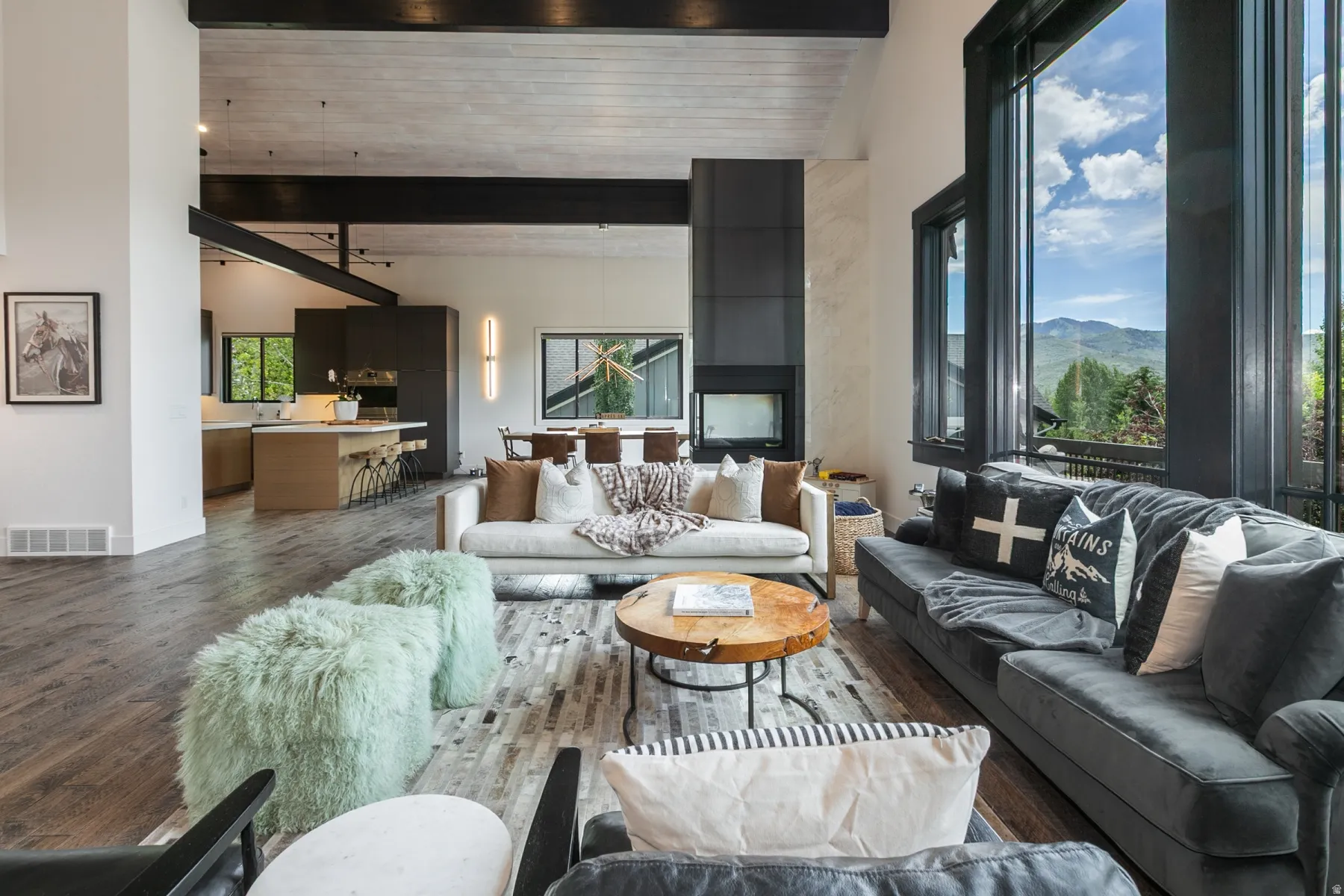 Spacious living area featuring large windows, wood-finish flooring, a modern fireplace, vaulted ceilings with exposed beams, and an open-concept layout