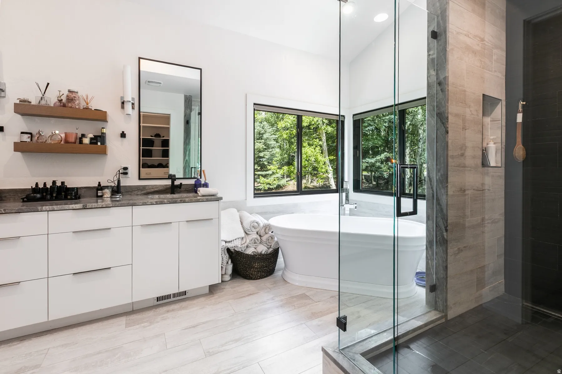 Modern bathroom with a freestanding soaking tub, expansive windows, glass-enclosed shower, stone-finish vanity countertop, and light wood-finish flooring