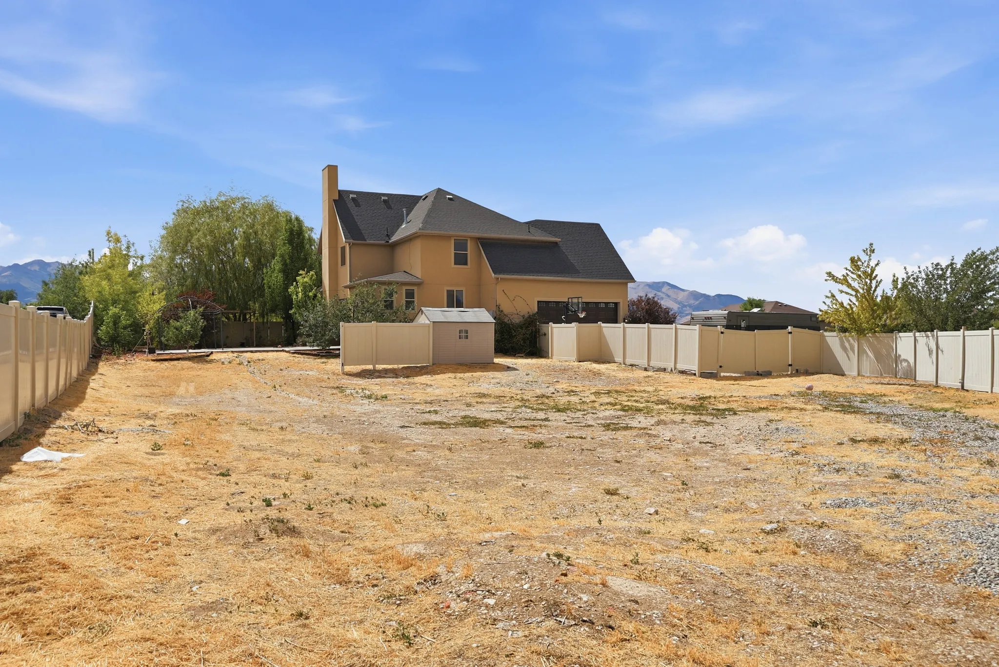 Expansive lot featuring a tan vinyl perimeter fence, a detached shed, mountain scenery, and a two-story residence in the background