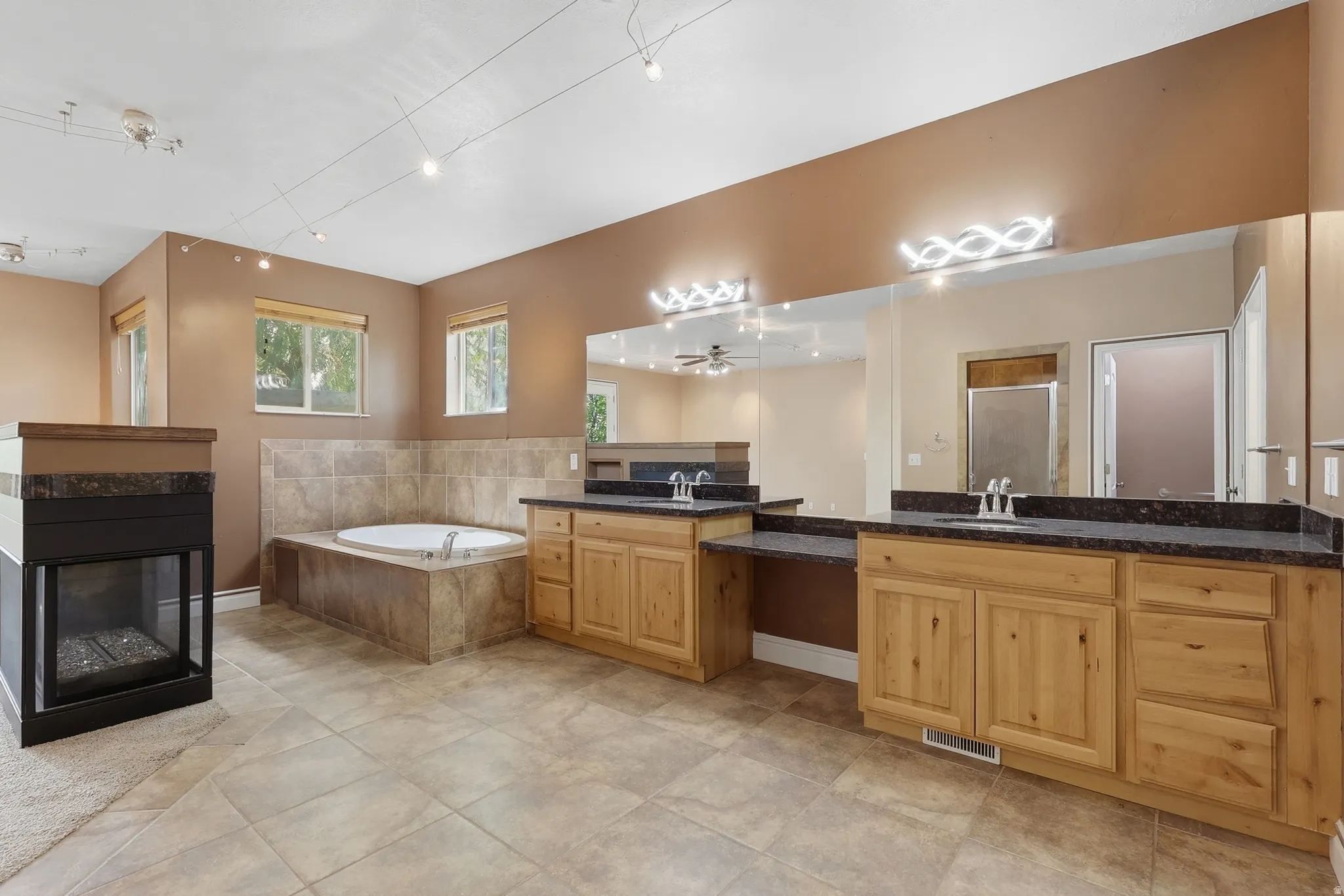 Spacious bathroom featuring dual vanities with dark countertops, a large soaking tub with tile surround, a built-in fireplace, tile flooring, and track lighting