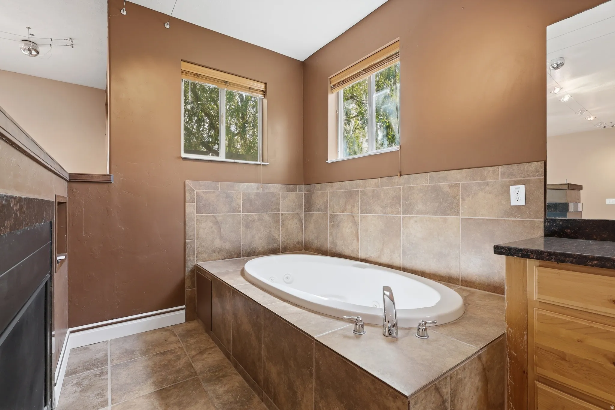 Soaking tub with a tiled surround, two windows, track lighting, a wood vanity, and a dark countertop