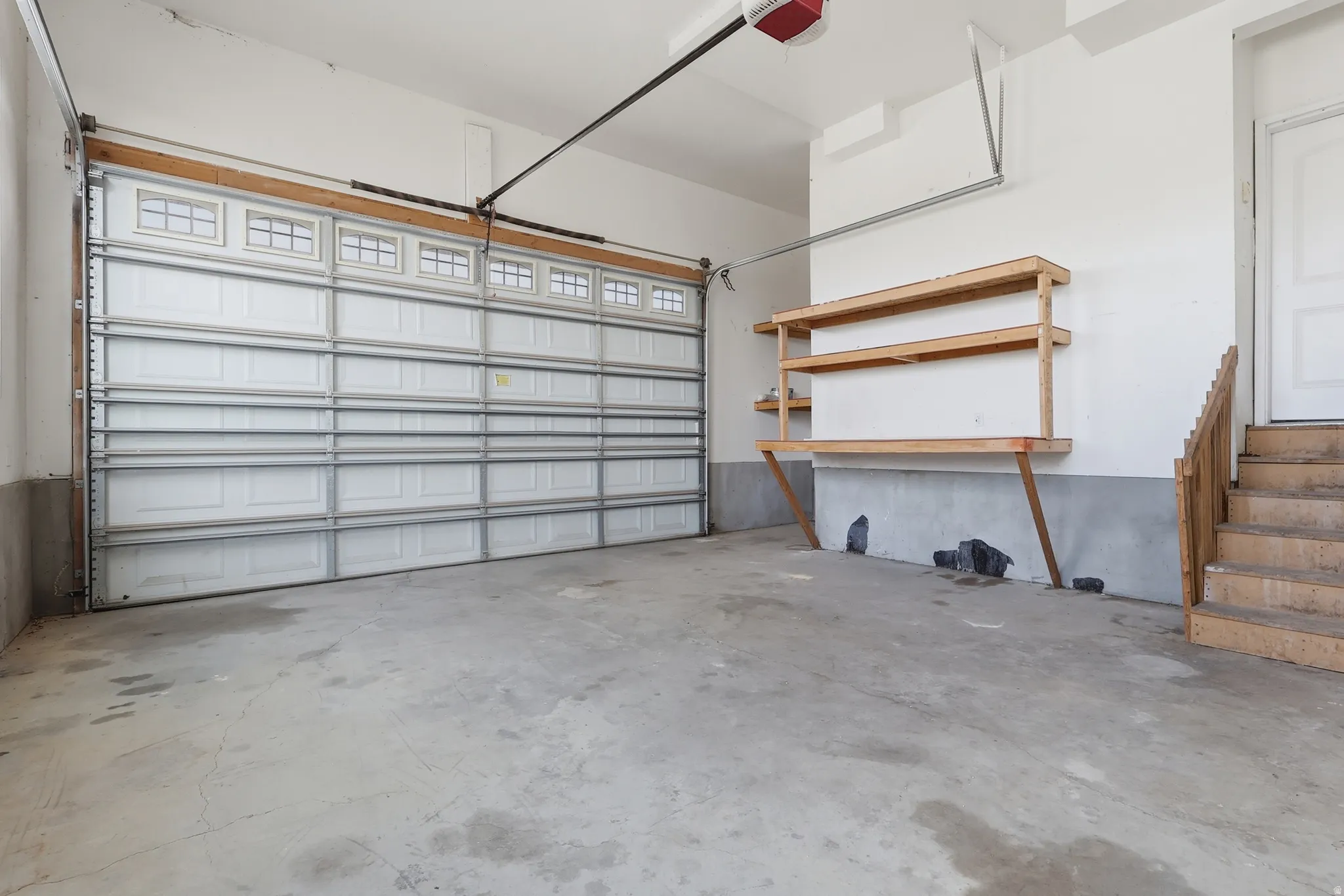 Spacious garage featuring a paneled overhead door with upper window inserts, concrete flooring, built-in wooden shelving, and a wooden staircase providing interior access