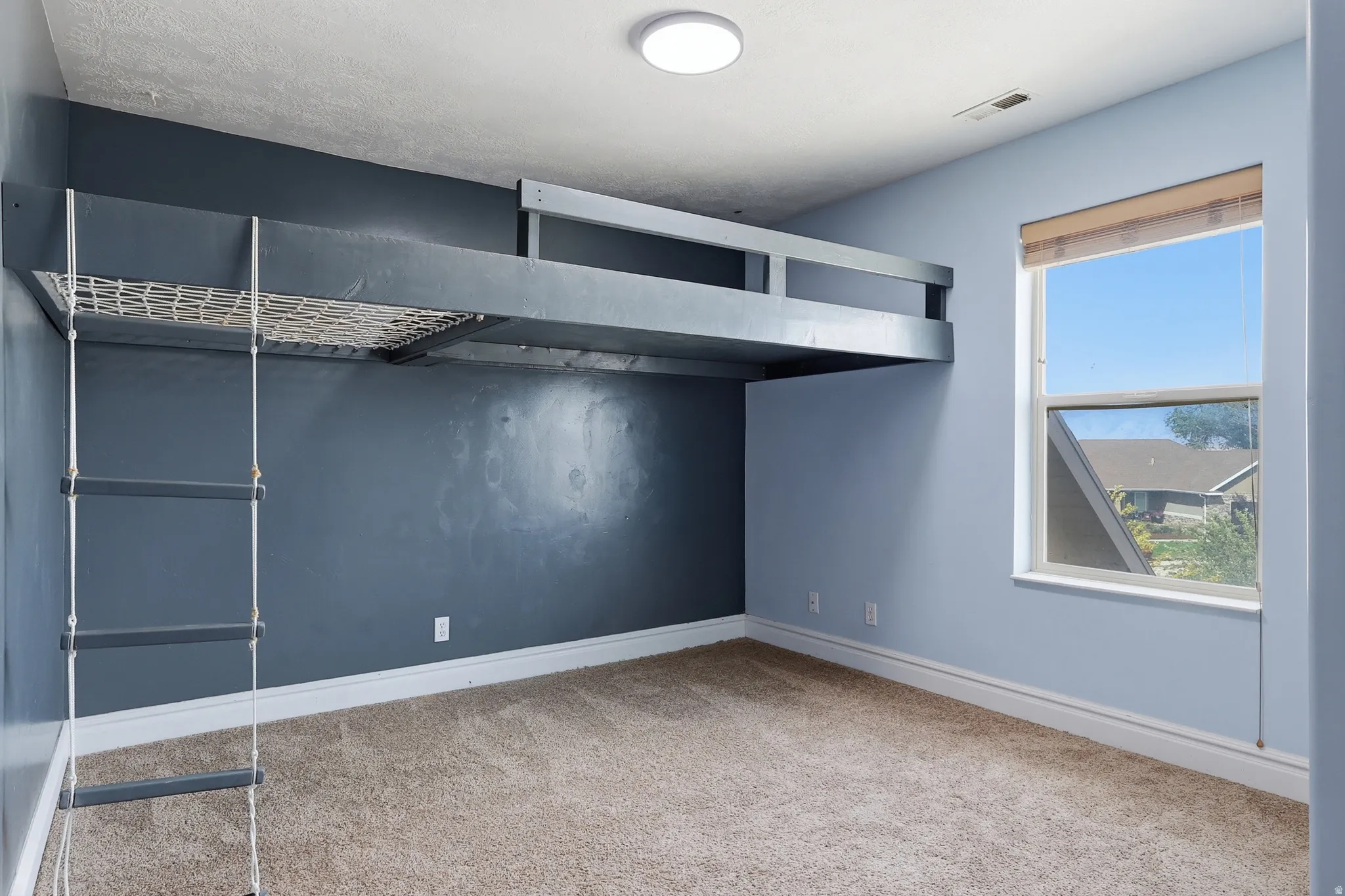 Built-in loft structure with rope ladder, light blue and charcoal accent walls, neutral carpet flooring, a single window with roller shade, and overhead flush-mount lighting