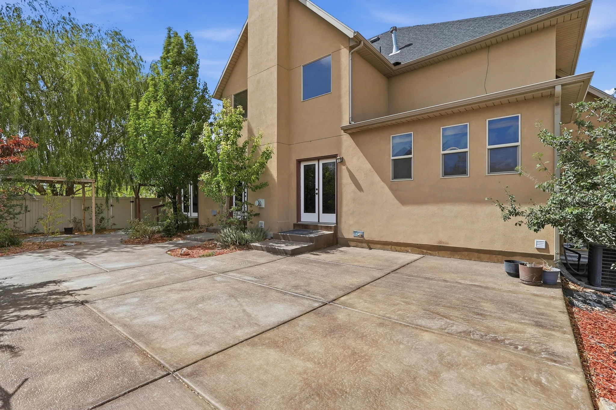 Spacious concrete patio featuring a stucco exterior, double glass French doors, multiple windows, and mature landscaping