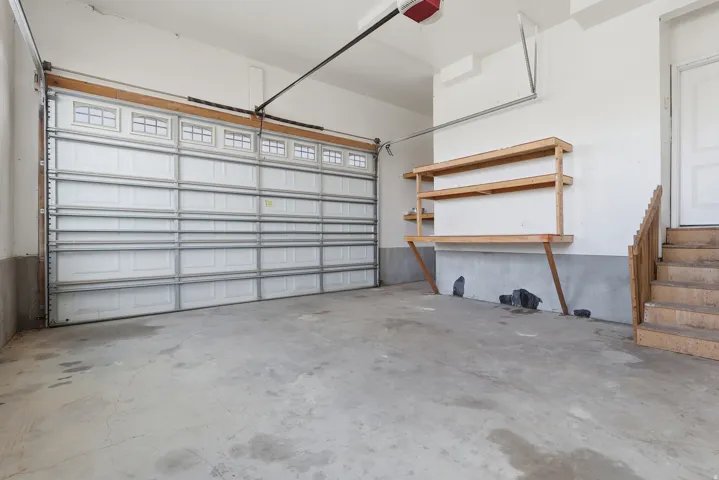 Spacious garage featuring a paneled overhead door with upper window inserts, concrete flooring, built-in wooden shelving, and a wooden staircase providing interior access