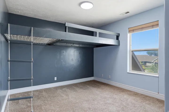 Built-in loft structure with rope ladder, light blue and charcoal accent walls, neutral carpet flooring, a single window with roller shade, and overhead flush-mount lighting