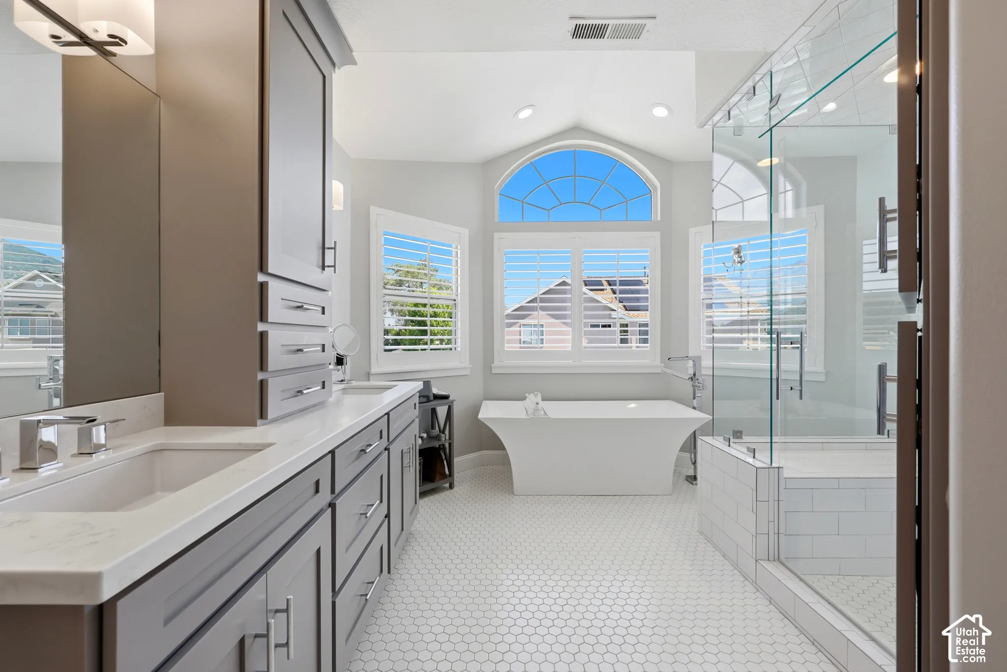 Vaulted ceiling bathroom featuring a freestanding tub, glass-enclosed shower, dual vanities with integrated sinks, and white hexagonal floor tiling