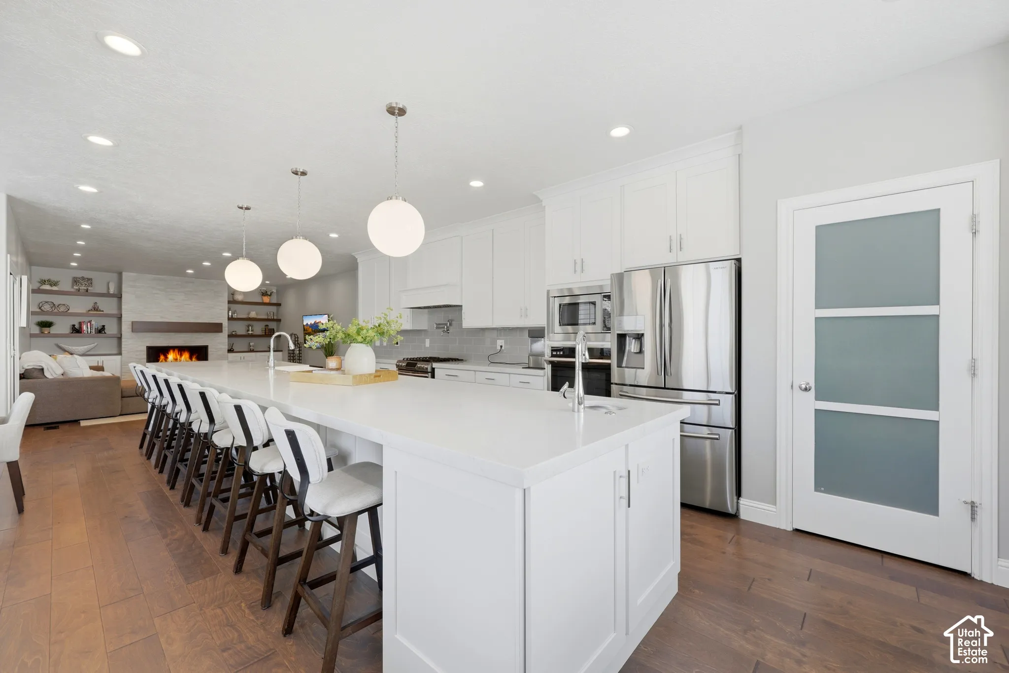 Open-concept kitchen and living area featuring an oversized island with seating, pendant lighting, stainless steel appliances, wood-finish flooring, and a linear fireplace