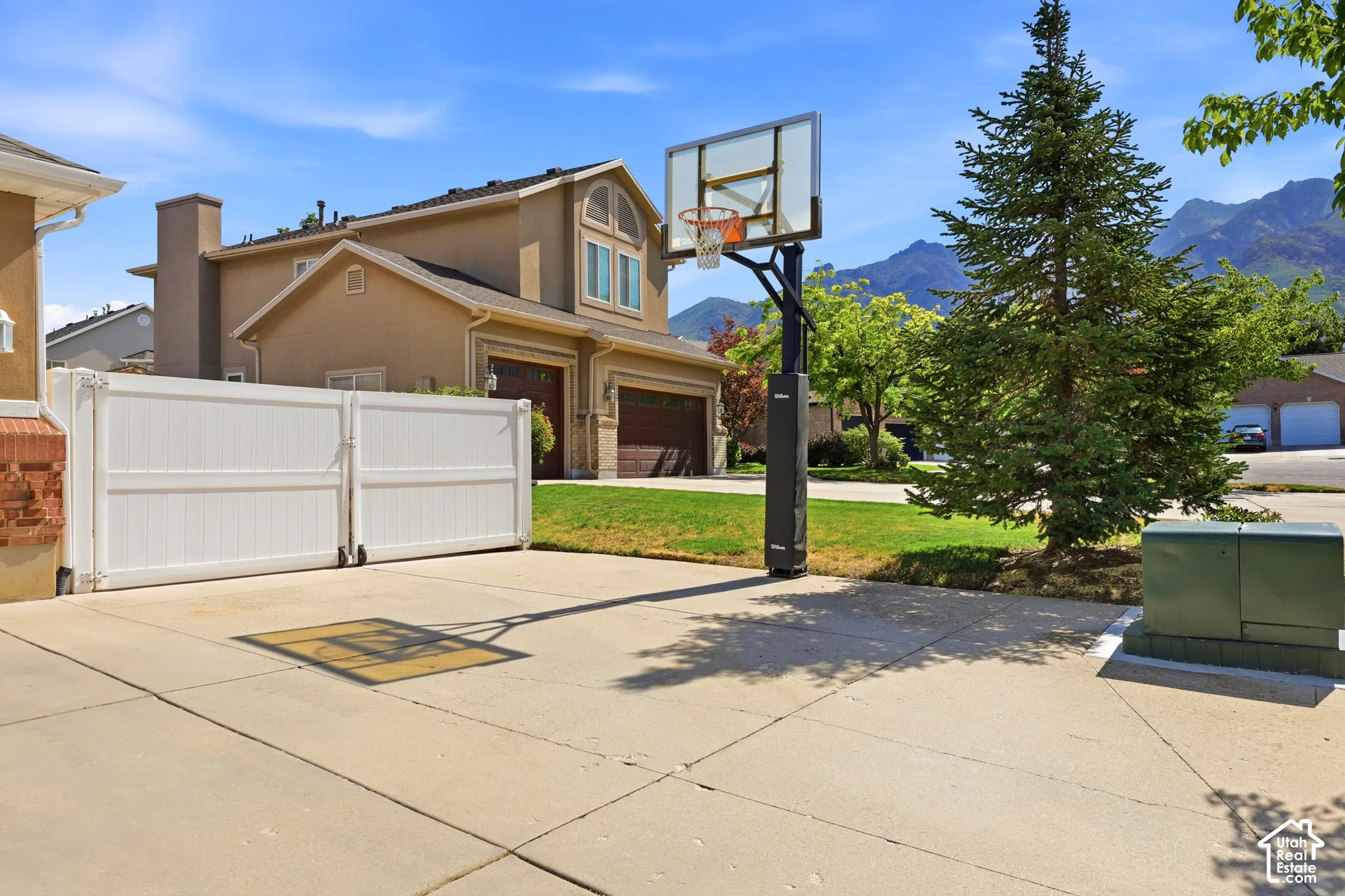 Concrete driveway with a movable basketball hoop, white privacy gate, and a mature evergreen tree