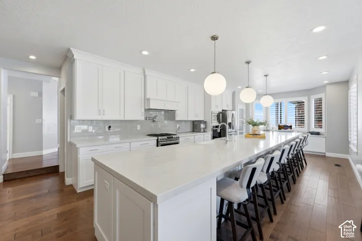 Spacious kitchen featuring an extended island with a light-toned countertop, white cabinetry with crown molding, a tiled backsplash, recessed lighting, and wood-finish flooring