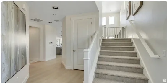 Carpeted staircase with white railings, featuring recessed lighting, light wood-finish flooring, a white paneled door, and a thermostat. PICTURE IS OF A SIMILAR HOME