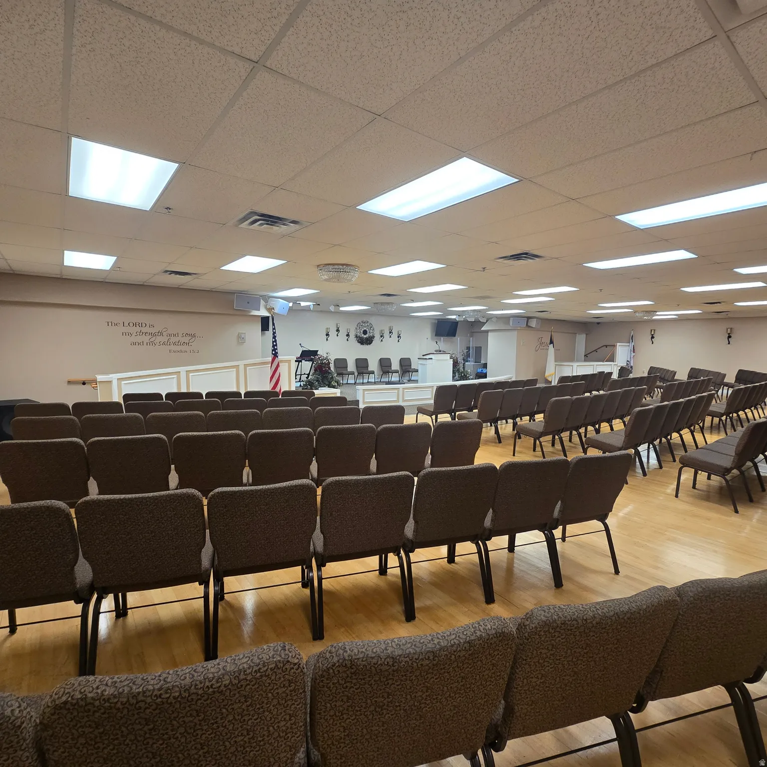 Spacious assembly hall featuring wood-finish flooring, drop ceiling with integrated lighting, and a raised platform stage