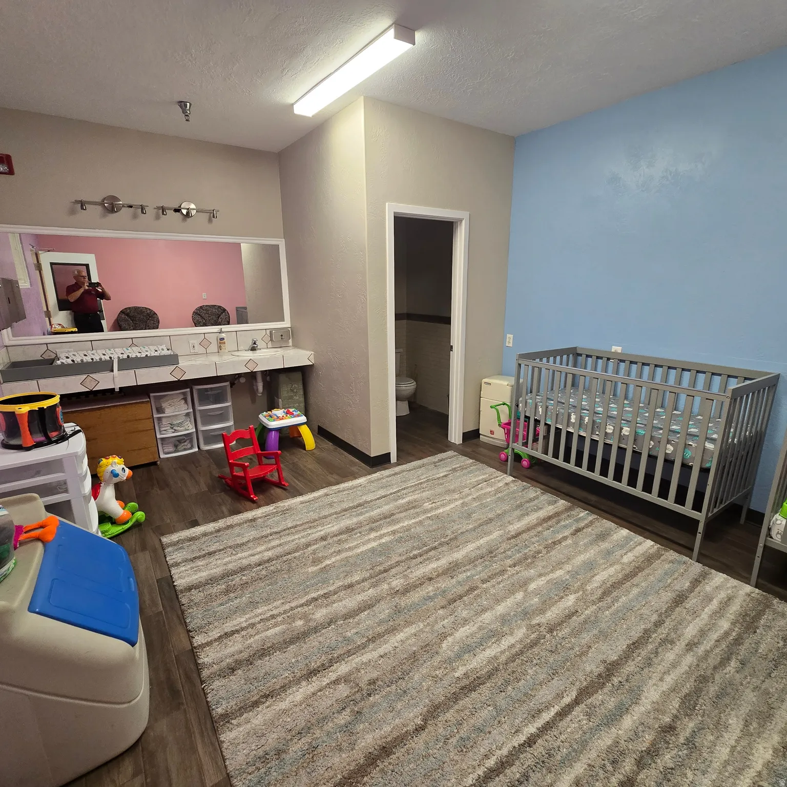 Spacious nursery room featuring wood-finish flooring, a large area rug, and dual vanity stations with tiled countertops. Toilet in closed area.