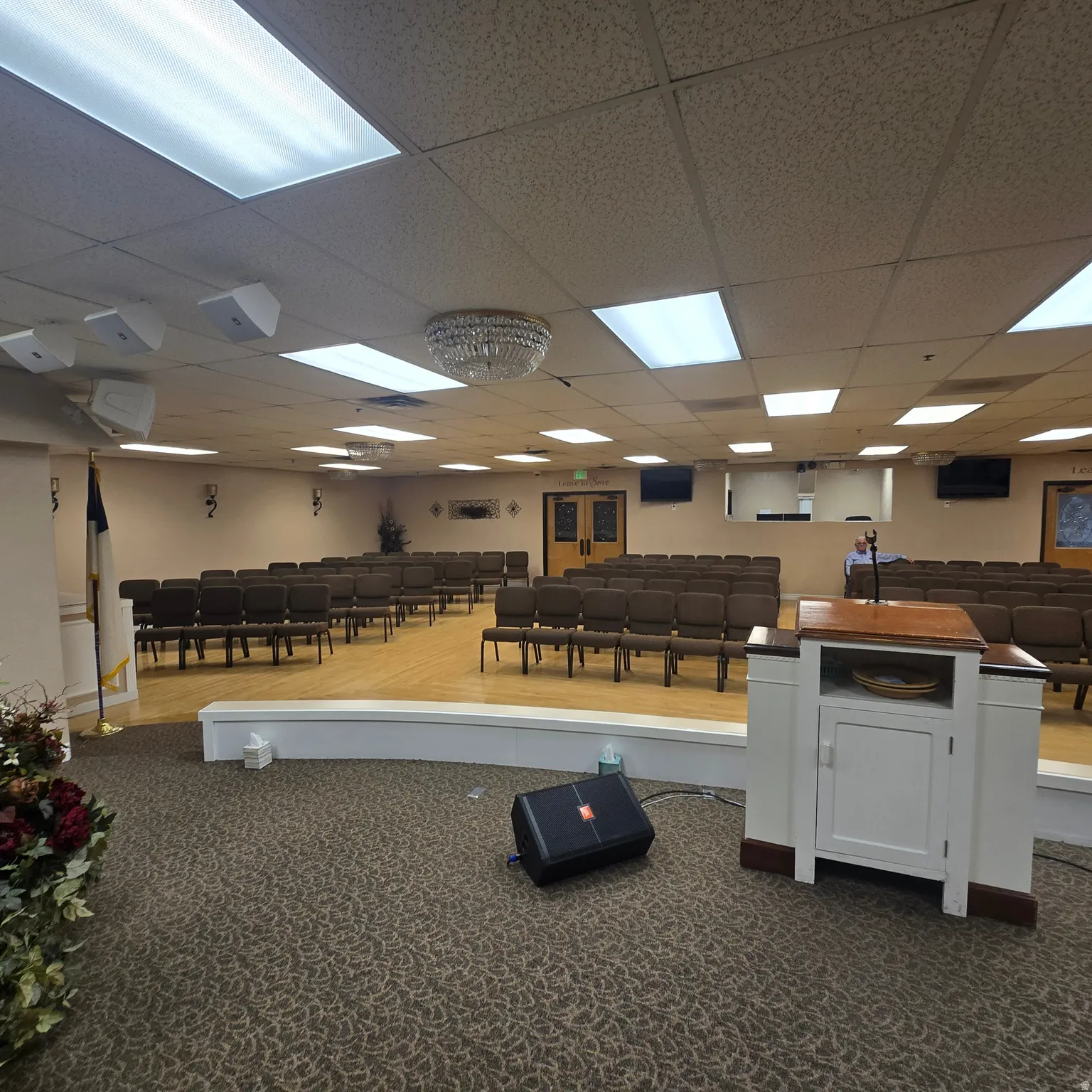 Spacious assembly room featuring a raised stage with wood-finish flooring, recessed fluorescent lighting, and a decorative central chandelier
