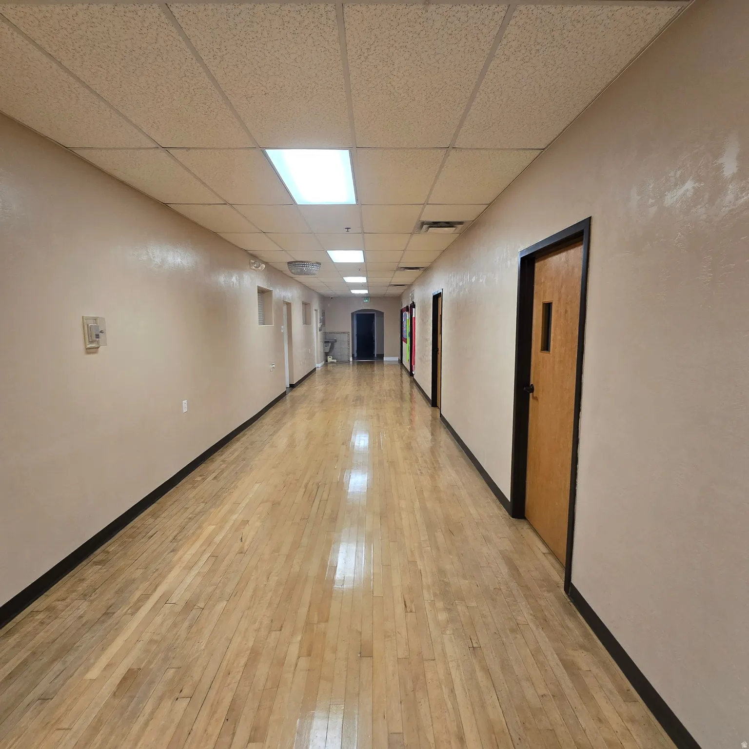 Long hallway featuring wood-finish flooring, drop ceiling with fluorescent lighting, and multiple entryways with wood doors
