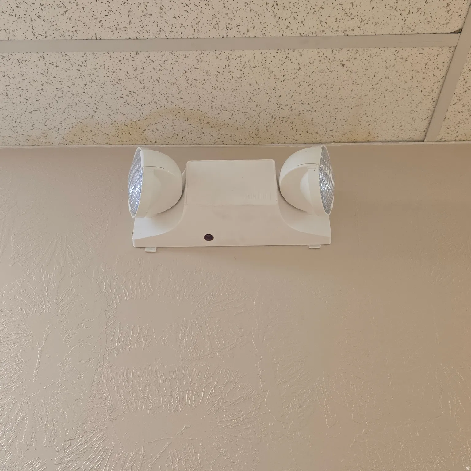 Wall-mounted emergency lighting fixture with dual adjustable heads, positioned below a suspended acoustic tile ceiling