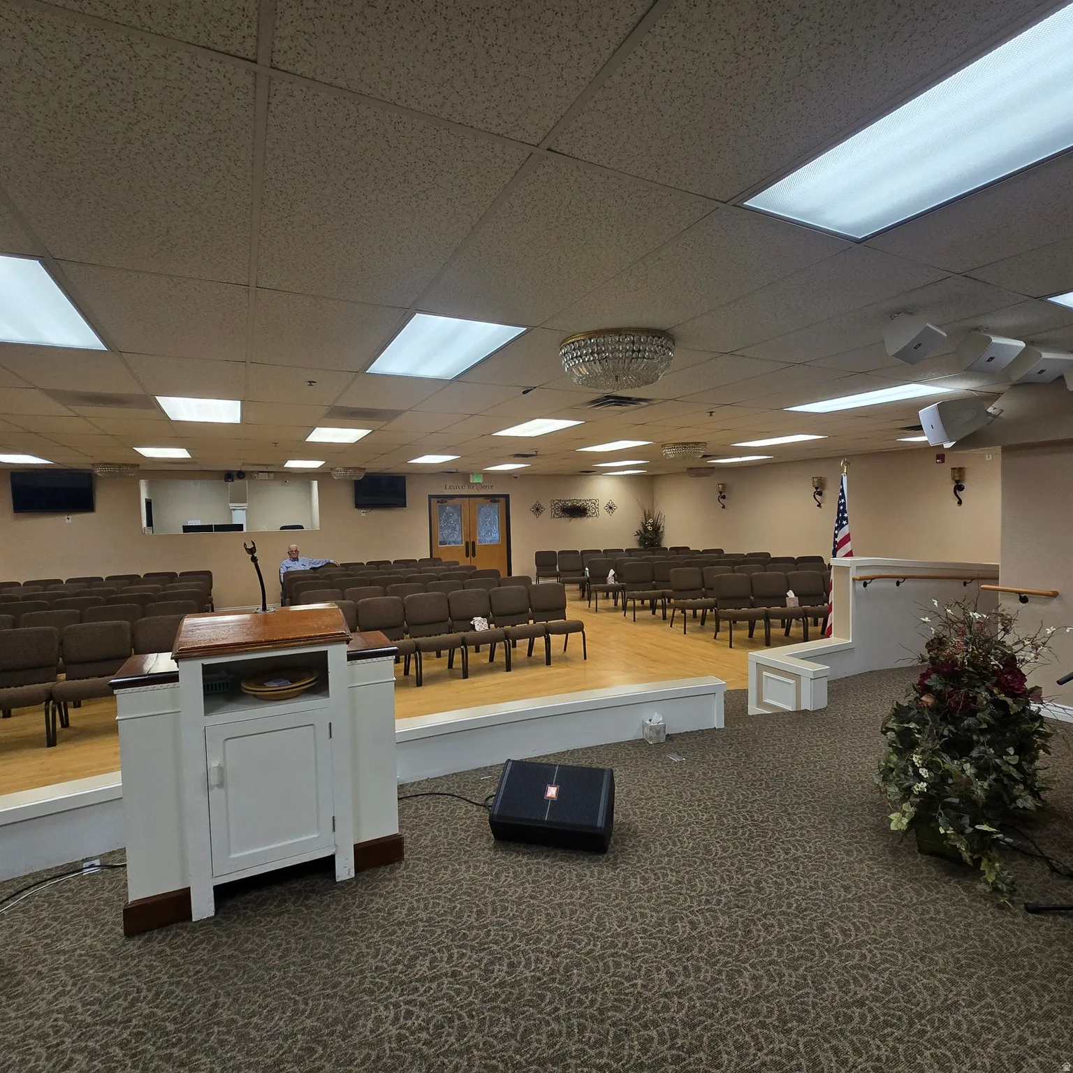 Spacious assembly room featuring a raised platform with wood-finish flooring, recessed fluorescent lighting, and a textured drop ceiling