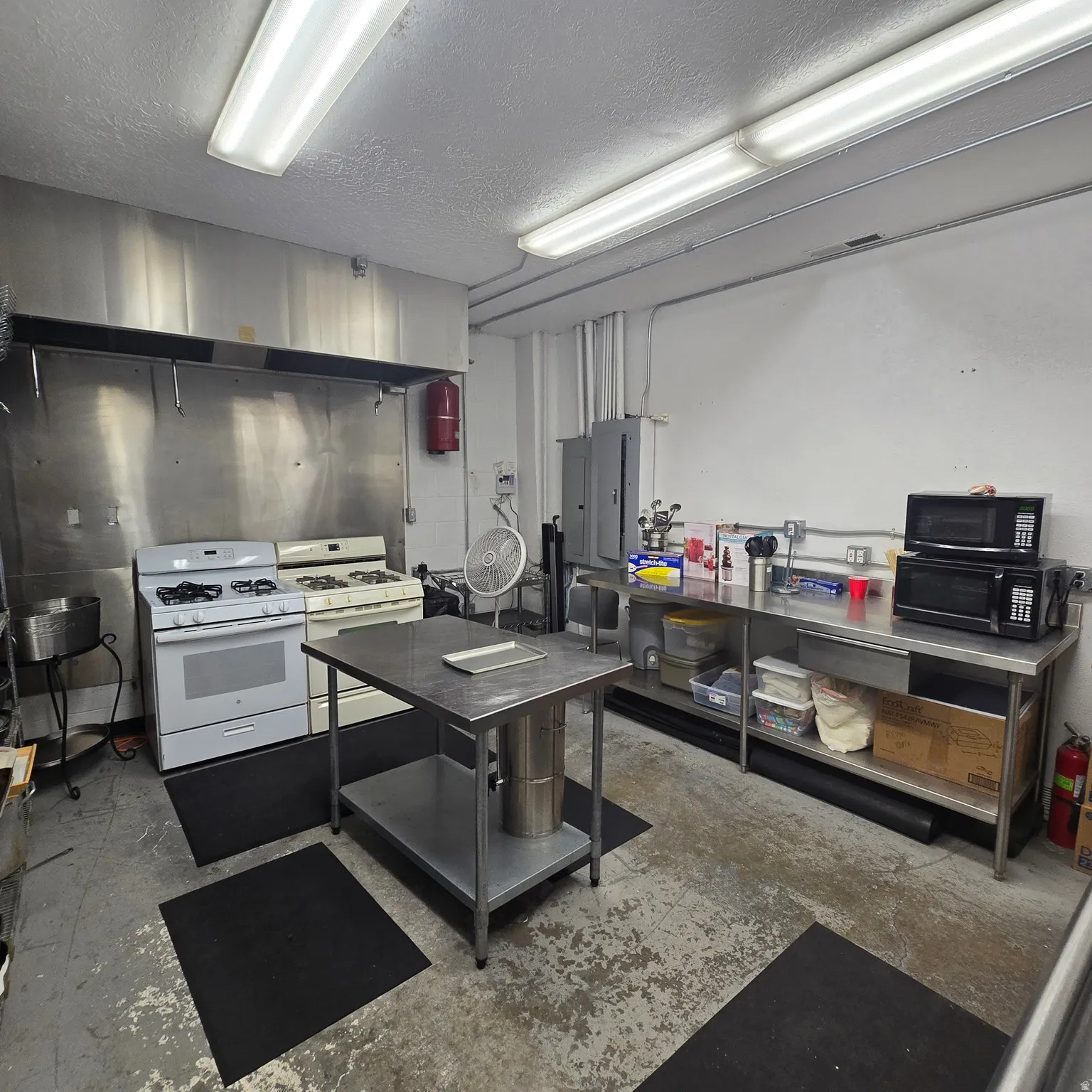 Commercial kitchen space featuring two gas ranges with a stainless steel vent hood, extensive stainless steel countertops, and a central stainless steel prep table