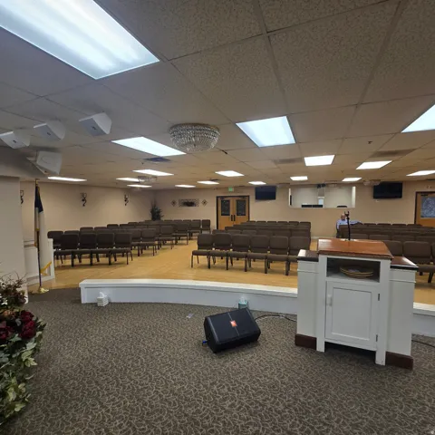 Spacious assembly room featuring a raised stage with wood-finish flooring, recessed fluorescent lighting, and a decorative central chandelier