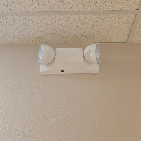 Wall-mounted emergency lighting fixture with dual adjustable heads, positioned below a suspended acoustic tile ceiling