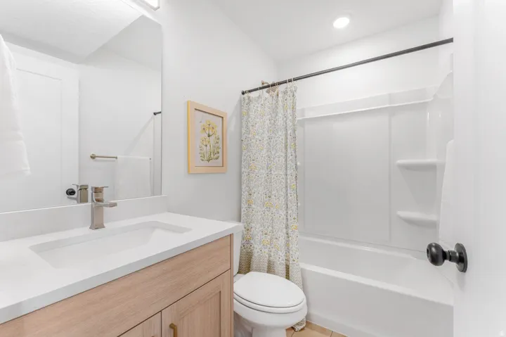 Bathroom featuring a light wood-finish vanity with a white countertop and undermount sink, a chrome faucet, a rectangular mirror, and a white tub/shower combination with an integrated shelving unit