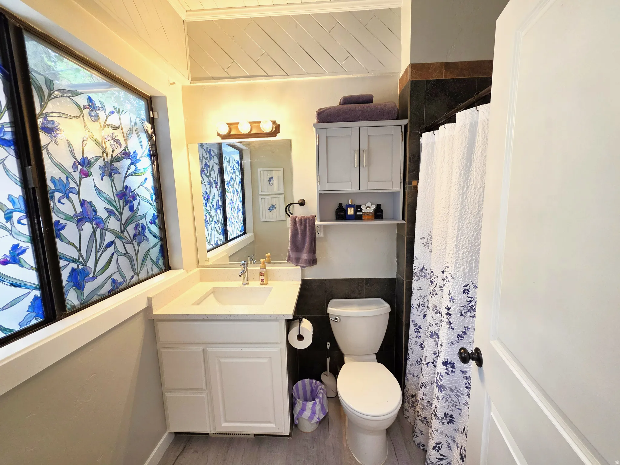 Bathroom featuring a single vanity with an integrated sink, decorative window film, patterned shower curtain, and an overhead storage cabinet