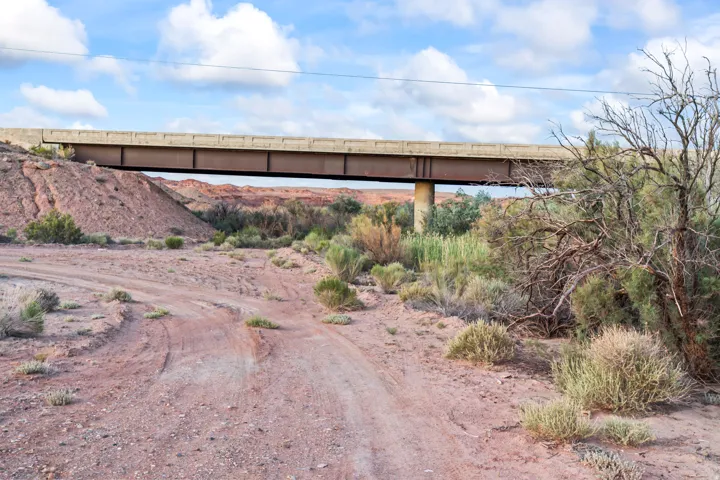 Unpaved road with visible tire tracks, surrounded by arid vegetation and a distant concrete and steel bridge structure