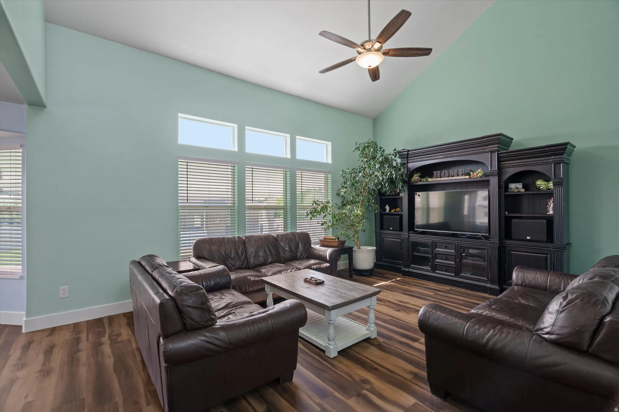 Vaulted ceiling living area featuring wood-finish flooring, a contemporary ceiling fan with integrated lighting, and multiple windows with horizontal blinds
