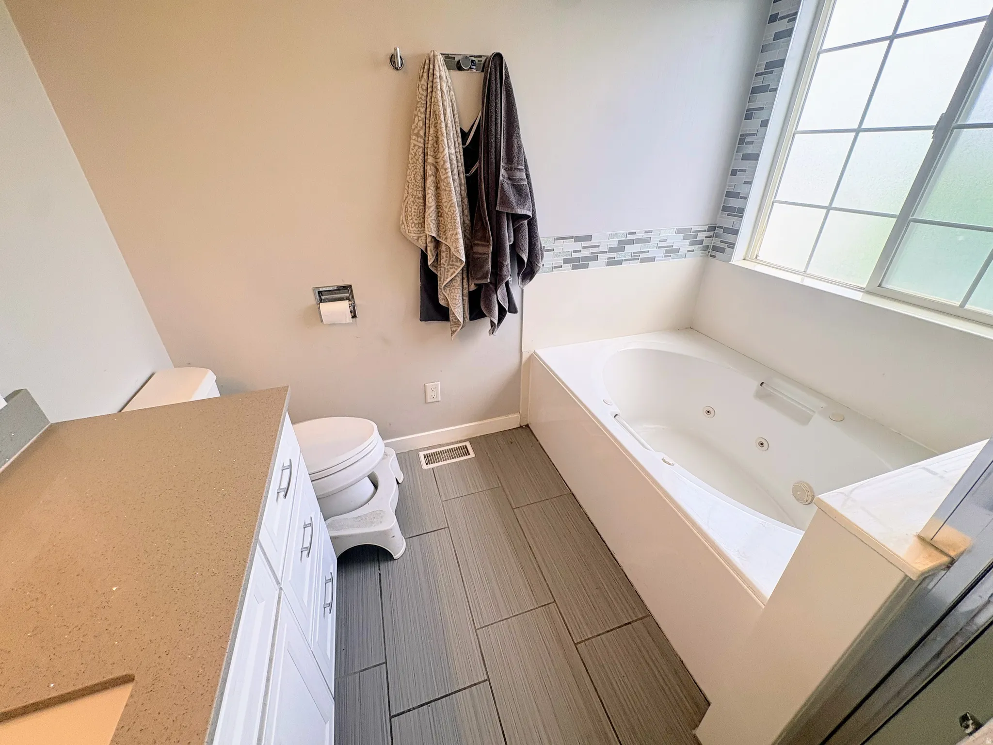 Bathroom featuring a jetted bathtub, a large window with privacy glass, mosaic tile detailing, a vanity with white cabinetry and a solid surface countertop, and wood-finish tile flooring