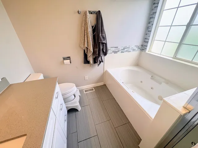 Bathroom featuring a jetted bathtub, a large window with privacy glass, mosaic tile detailing, a vanity with white cabinetry and a solid surface countertop, and wood-finish tile flooring