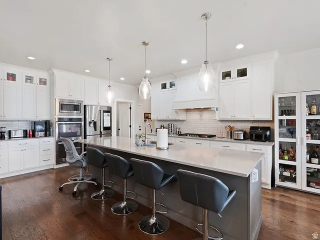 Spacious kitchen featuring a large central island with seating, stainless steel appliances, white cabinetry with glass-front accents, dark wood-finish flooring, and a white subway tile backsplash