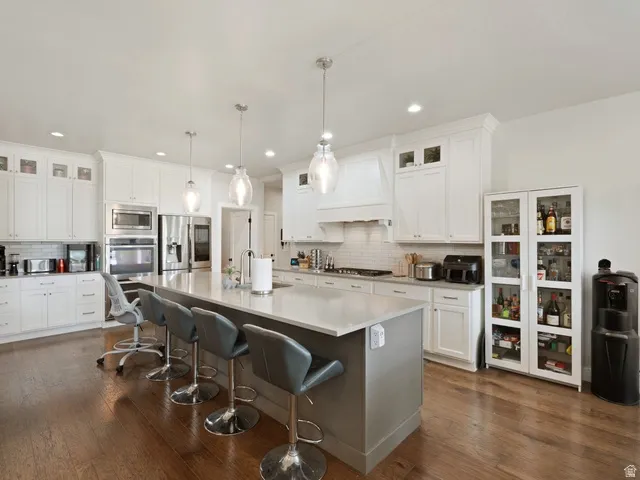 Gourmet kitchen featuring a substantial central island with seating, white shaker-style cabinetry, stainless steel appliances, pendant lighting, and wood-finish flooring