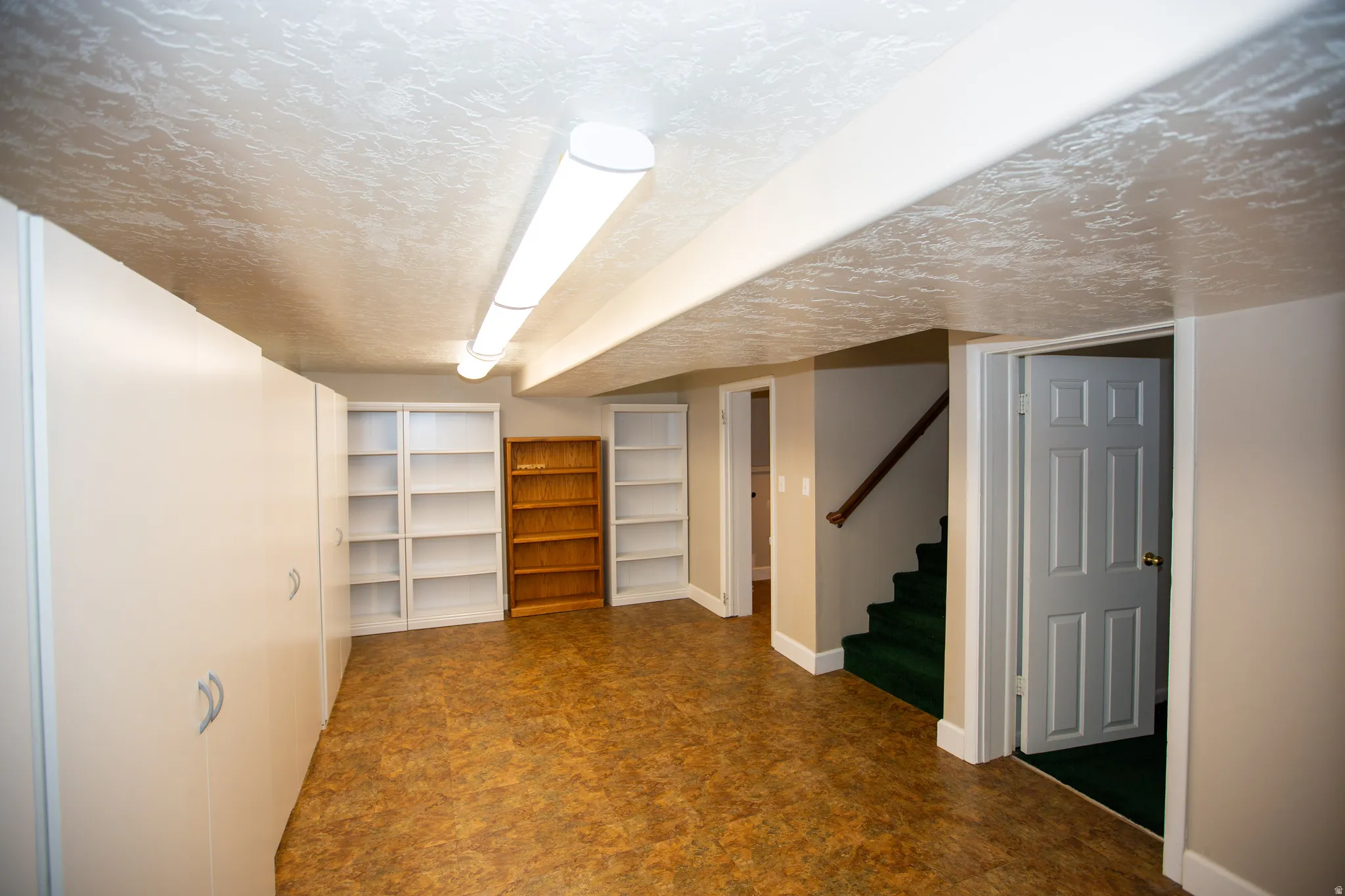 Spacious multi-purpose room featuring extensive built-in storage cabinetry, multiple wall-mounted bookshelves, wood-finish flooring, and bright overhead lighting