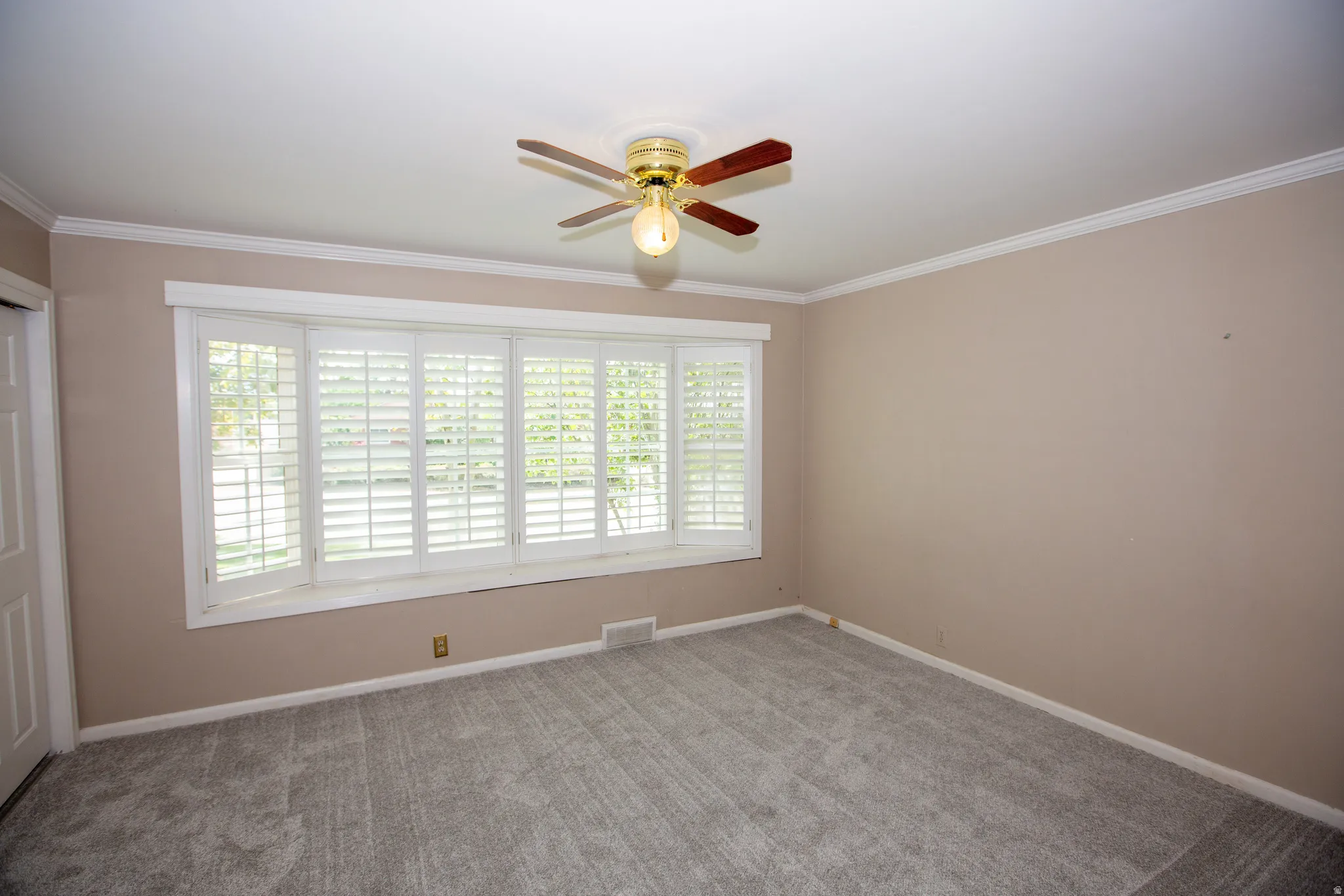 Room featuring a prominent bay window with white plantation shutters, crown molding, a ceiling fan with light fixture, neutral wall paint, and gray carpet flooring