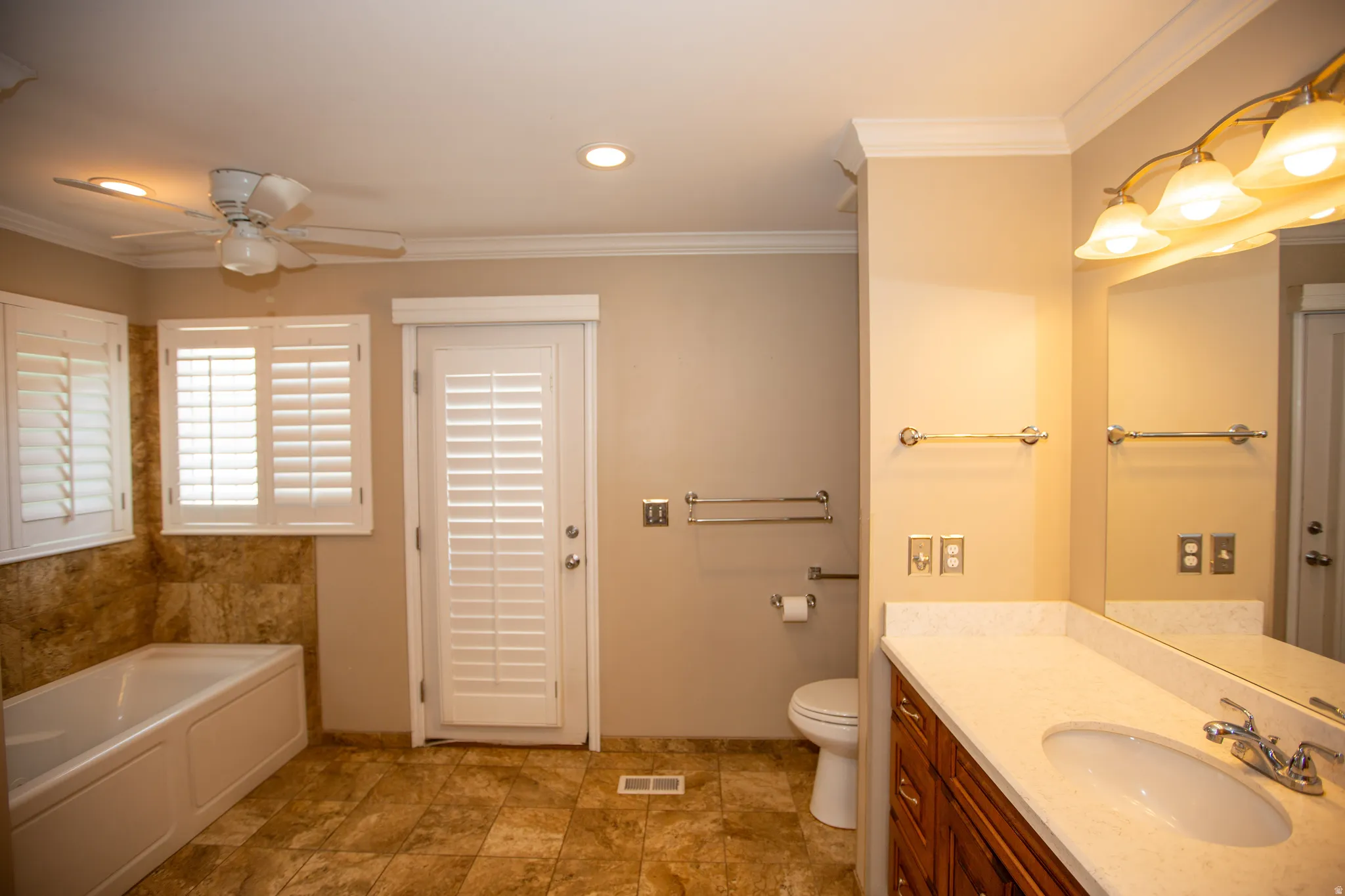 Primary bedroom bath featuring a built-in tub with tile surround, white plantation shutters, a ceiling fan, and a double vanity with a light-colored countertop, with ensuite laundry