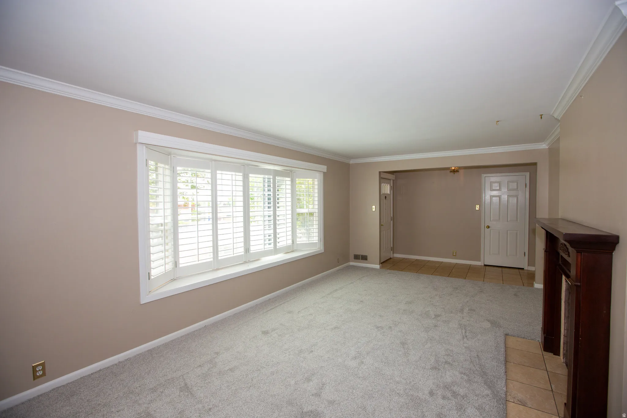 Living area with light gray carpeting, a prominent bay window with white plantation shutters, and crown molding