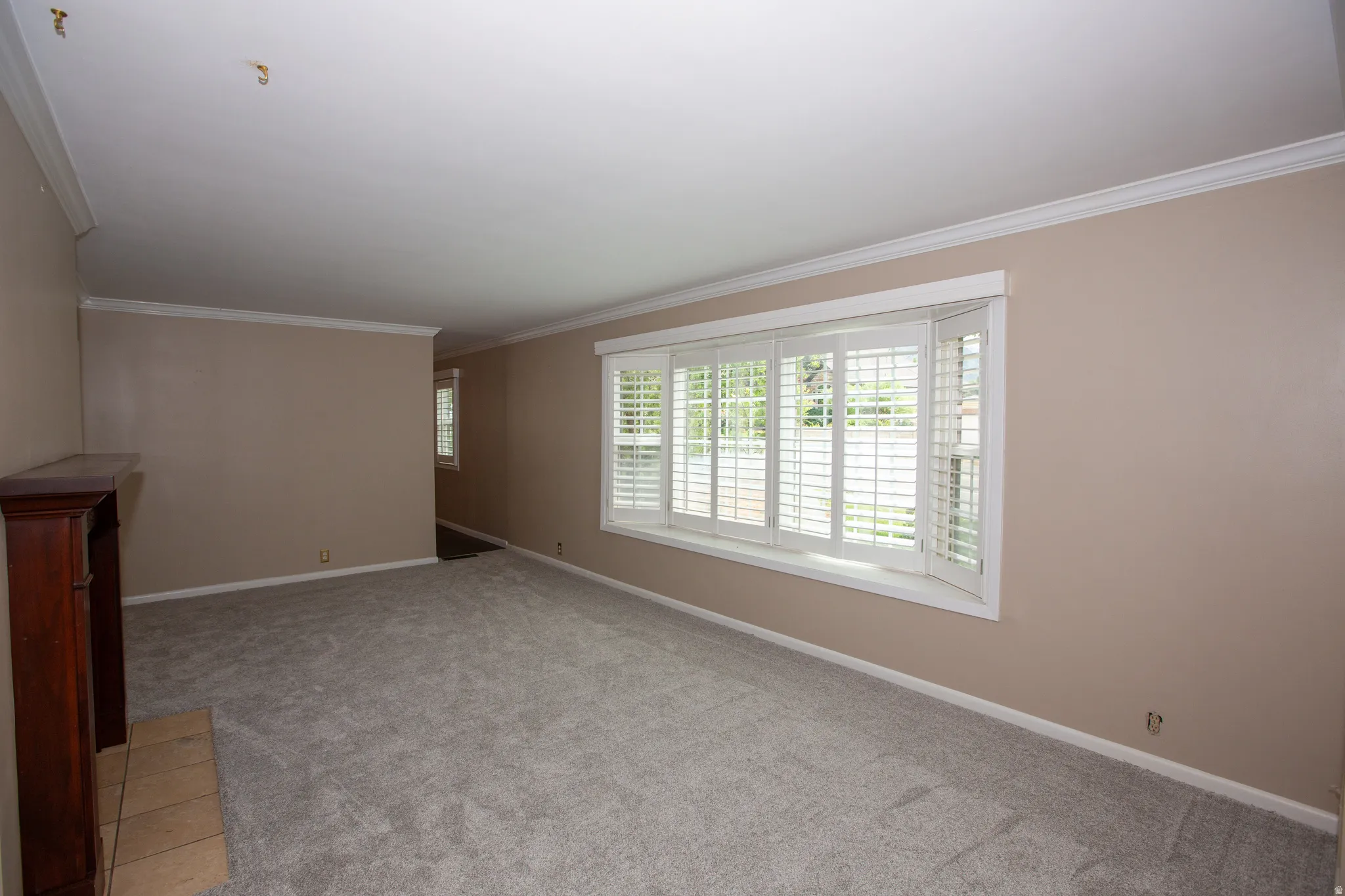 Spacious room featuring a bay window with plantation shutters, neutral wall tones, light gray carpeting, crown molding, and a fireplace with a wood mantel and tile hearth