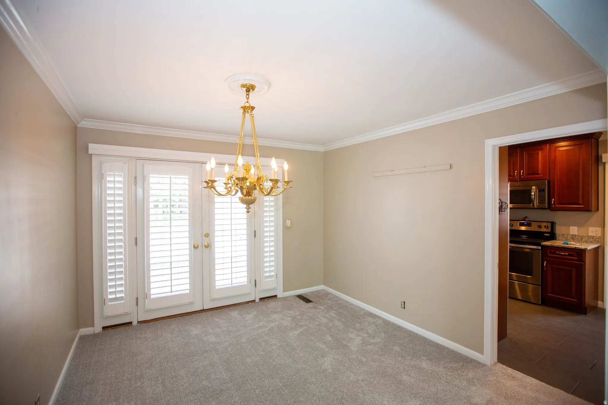 Carpeted room featuring a brass chandelier with ceiling medallion, crown molding, and white plantation shutters on French doors